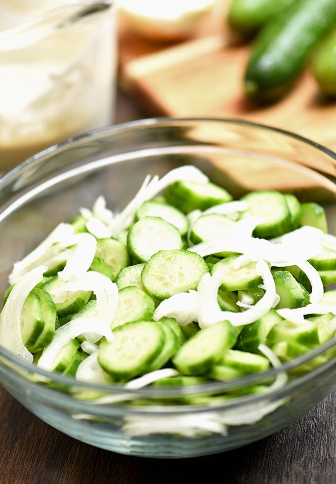 sliced cucumbers and sweet onions in a clear bowl with a side of mayo dressing