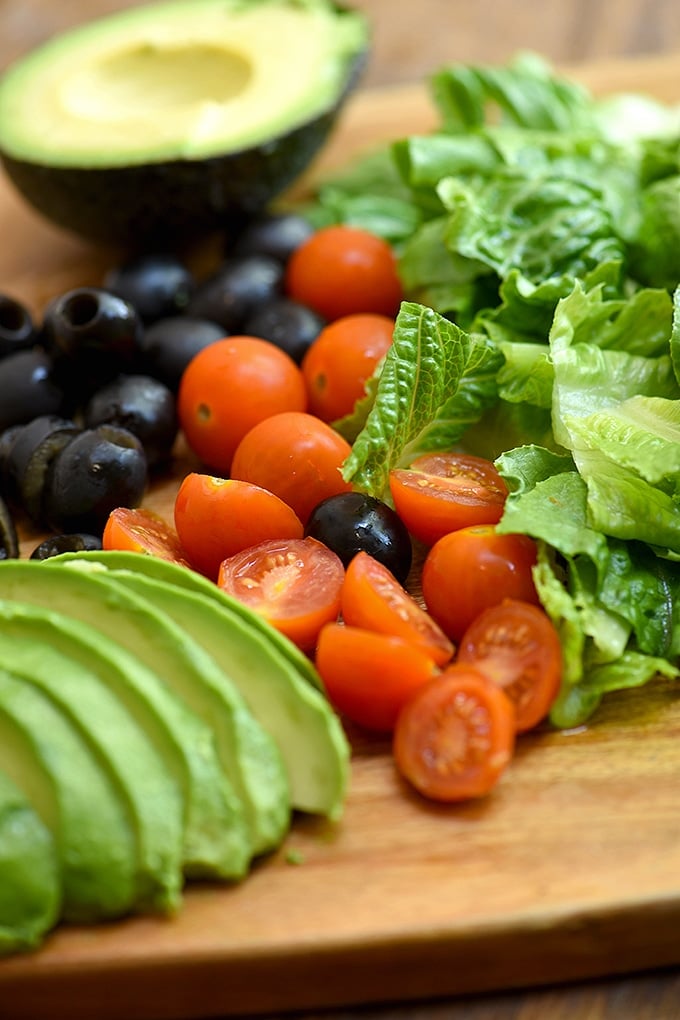 chopped romaine lettuce, cherry tomatoes, black olives, and sliced avocados on a wood cutting board