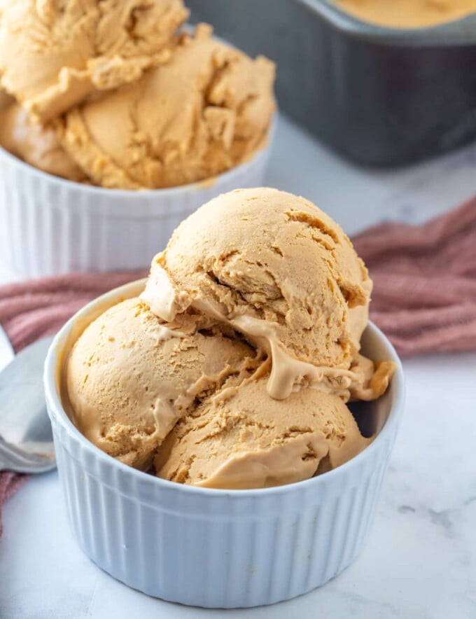 scoops of dulce de leche ice cream in white bowls with a metal loaf pan of ice cream in the background.