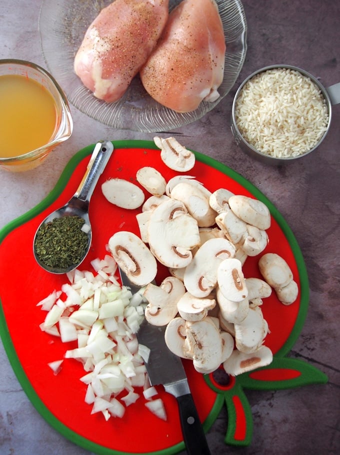 sliced mushrooms and onions on a cutting board, chicken breast, rice, and chicken broth on the side