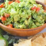 Homemade guacamole in brown bowl with chips, jalapenos, and avocado in background.