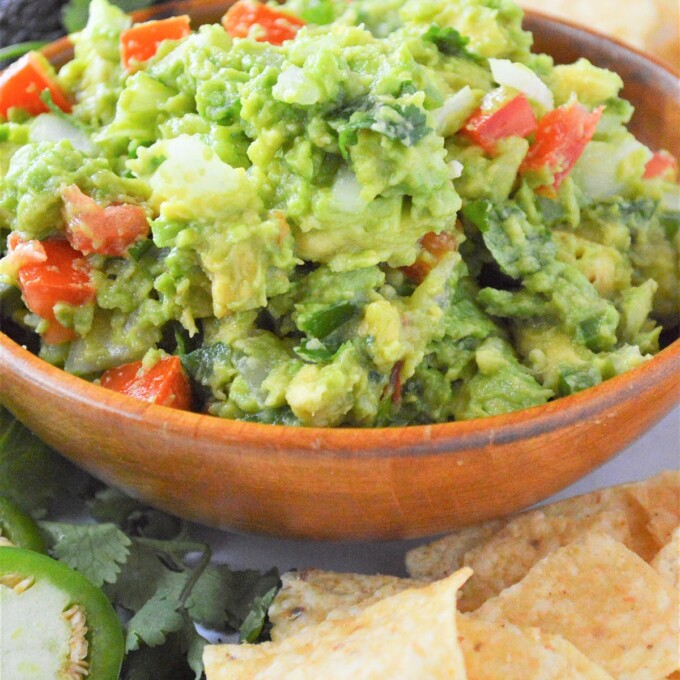 Homemade guacamole in brown bowl with chips, jalapenos, and avocado in background.