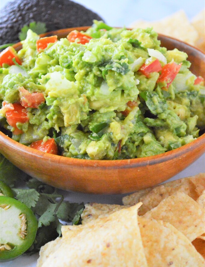 Homemade guacamole in brown bowl with chips, jalapenos, and avocado in background.
