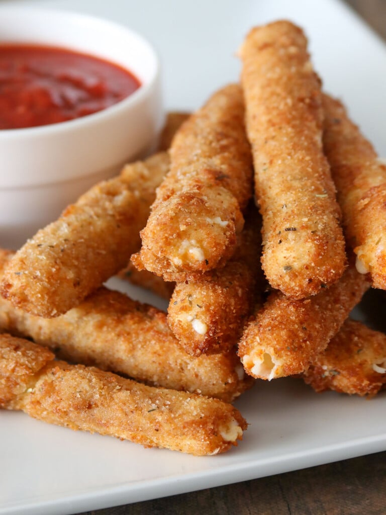 Homemade Mozzarella Sticks on a white plate with a bowl of Marinara Sauce on the side.