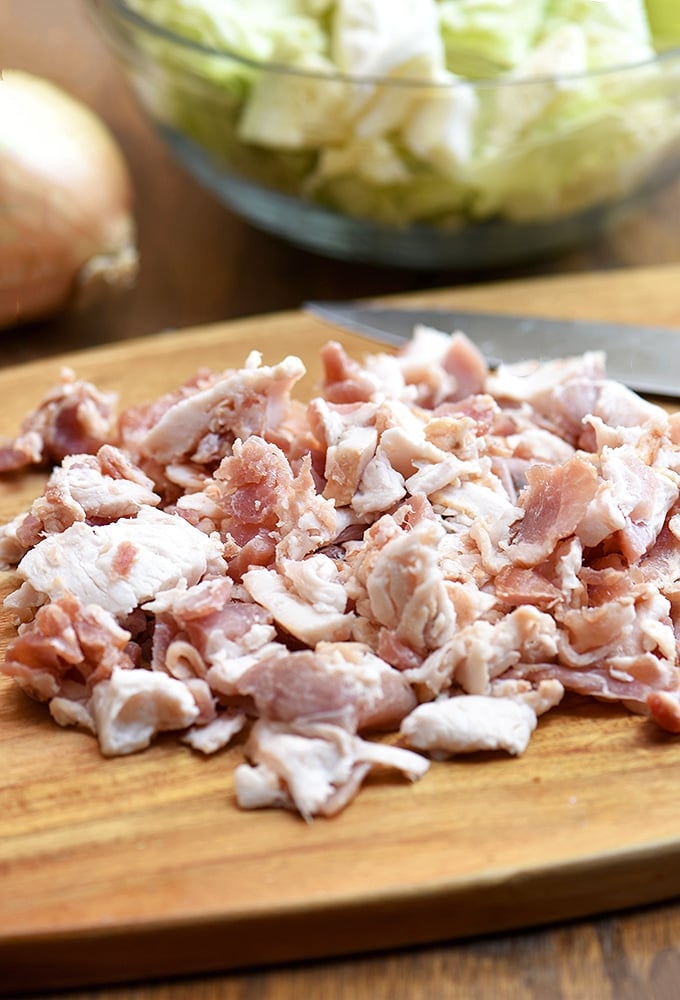 chopped bacon ends and pieces on a cutting board, a bowl of chopped cabbage, and a whole onion