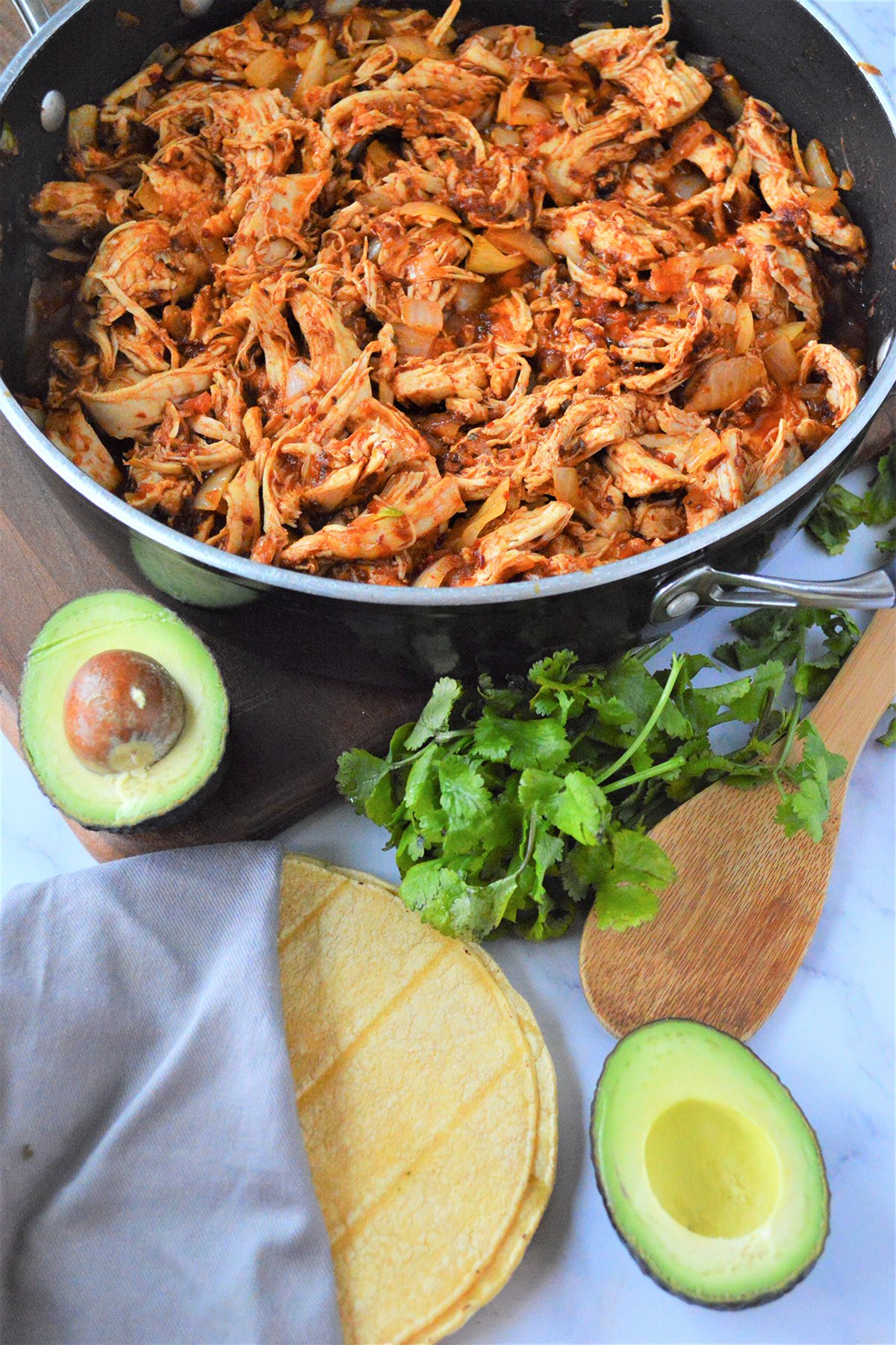Mexican Chicken Tinga in a pan with sliced avocados, corn tortilla,m and cilantro on the side