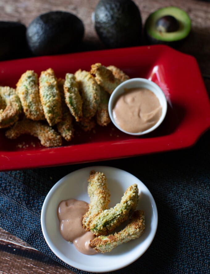 baked avocado fries in a white plate with dipping sauce with red platter with avocado fries on the side