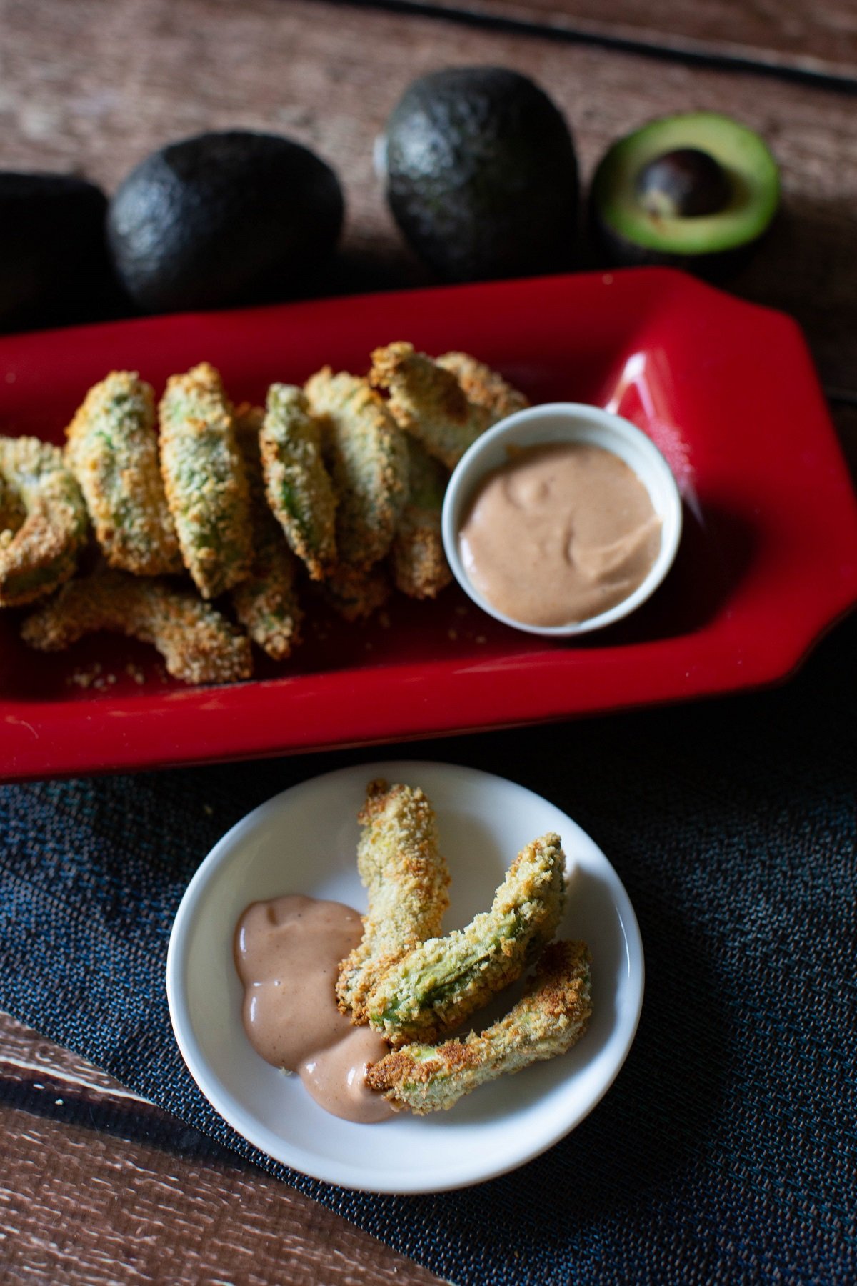 baked avocado fries in a white plate with dipping sauce with red platter with avocado fries on the side