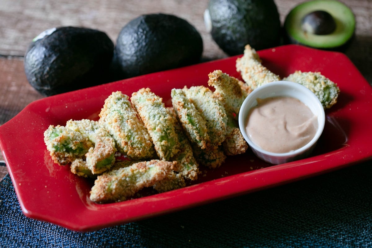 baked avocado fries and dipping sauce on a platter with avocados on the side