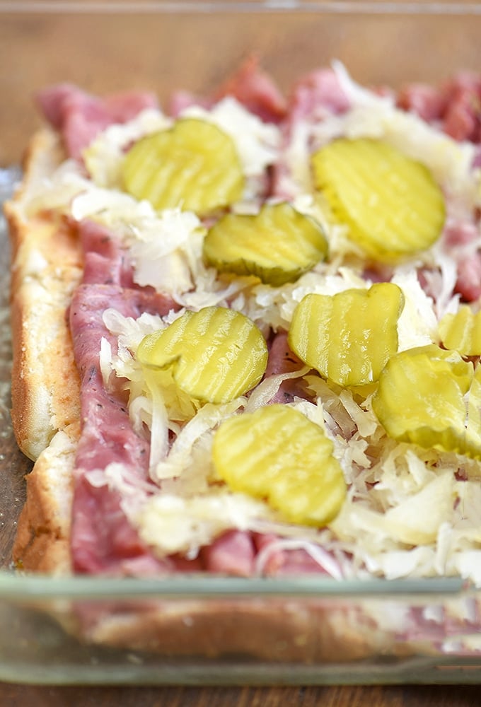 Hawaiian rolls topped with thousand island dressing, pastrami, sauerkraut and pickles in a baking dish.