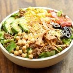 Taco Salad with lettuce, ground beef, avocados, cherry tomatoes, garbanzo beans, olives, tortilla strips and cheese in a white serving bowl