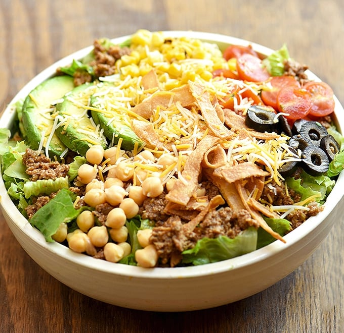 Taco Salad with lettuce, ground beef, avocados, cherry tomatoes, garbanzo beans, olives, tortilla strips and cheese in a white serving bowl