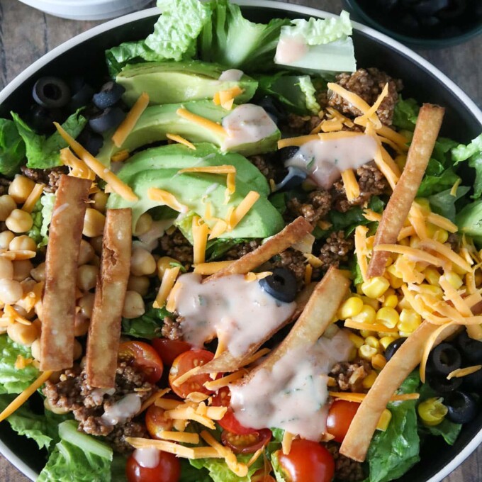 Taco Salad with lettuce, ground beef, avocados, cherry tomatoes, garbanzo beans, olives, tortilla strips and cheese in a serving bowl