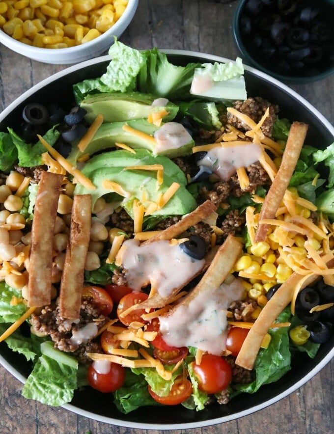 Taco Salad with lettuce, ground beef, avocados, cherry tomatoes, garbanzo beans, olives, tortilla strips and cheese in a serving bowl