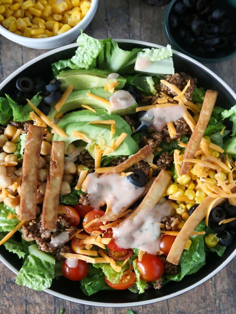 Taco Salad with lettuce, ground beef, avocados, cherry tomatoes, garbanzo beans, olives, tortilla strips and cheese in a serving bowl