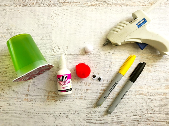 green Jello snack cups, red pom-pom, white pom-pom, googly eyes, black sharpie, yellow sharpie, and hot glue gun on a white board