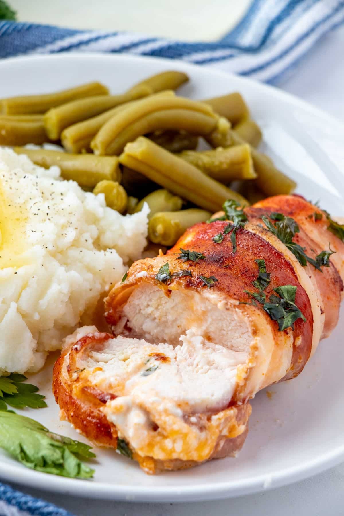 sliced bacon-wrapped cheese-stuffed chicken breast on a white plate with mashed potatoes and green beans on the side.