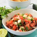 Homemade Pico de Gallo in a white serving bowl with tortilla chips in the background.