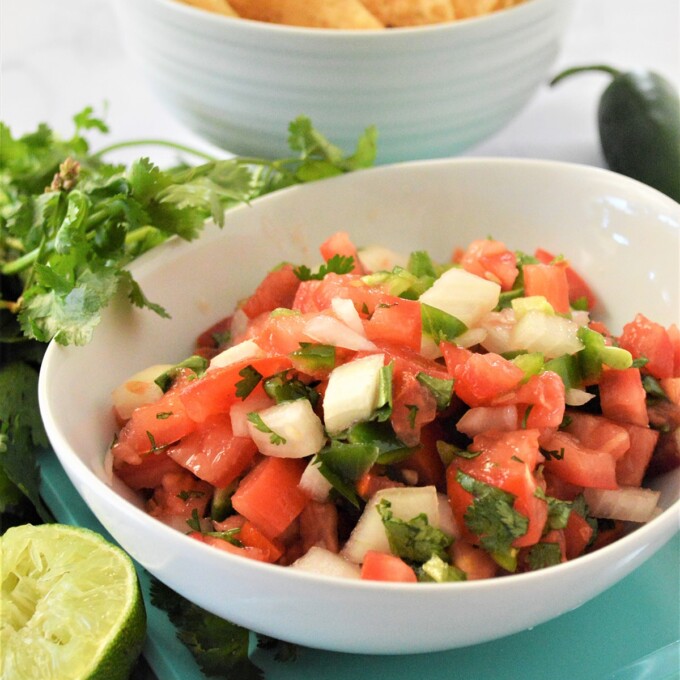 Homemade Pico de Gallo in a white serving bowl with tortilla chips in the background.