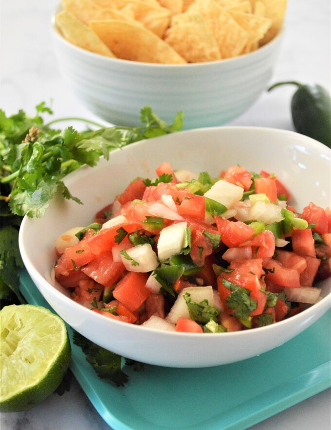 Homemade Pico de Gallo in a white serving bowl with tortilla chips in the background.